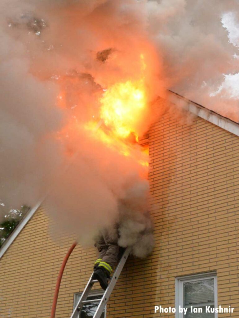 A firefighter with a hoseline on a ladder is engulfed by smoke and flames