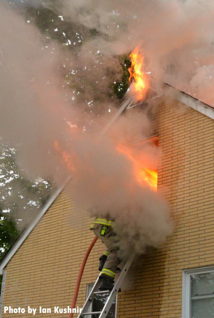 Flames roar from a window, obscuring a firefighter who has climbed a ladder