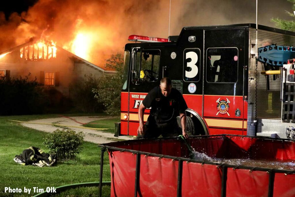 Portable tank being filled while flames roar through home in background