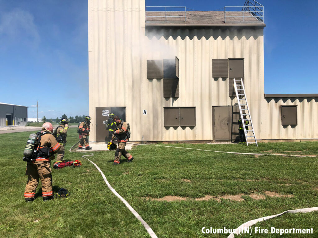 Smoke from the training building with a ladder thrown and firefighters with hoselines