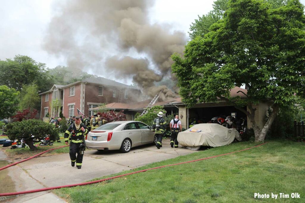 Two cars in the driveway of home with smoke showing and a ladder thrown on roof