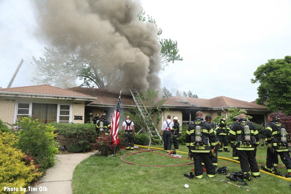 Firefighters in the yard at a ranch style home with a ladder and smoke showing