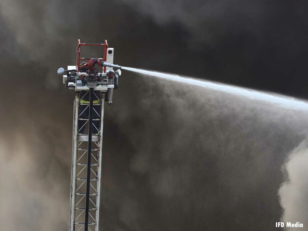 An elevated master stream on an aerial sprays water with smoke behind it