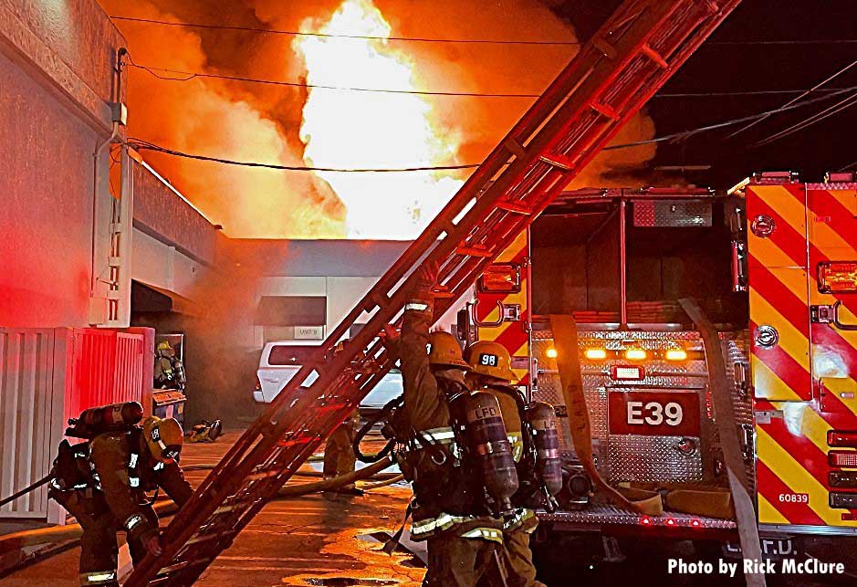 Firefighters raise a ladder as flames rage behind them and a fire truck