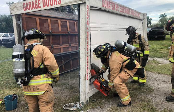 (5) One firefighter coaches another firefighter as he finishes one of his cuts on this sectional garage door.