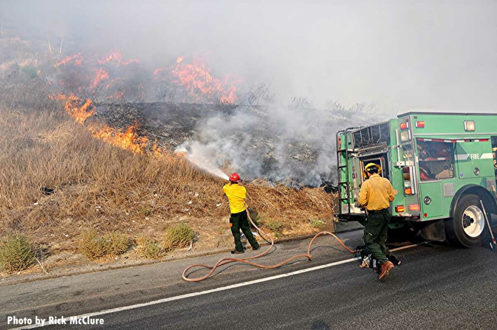 Firefighters train hoseline on wildfire flames on side of road