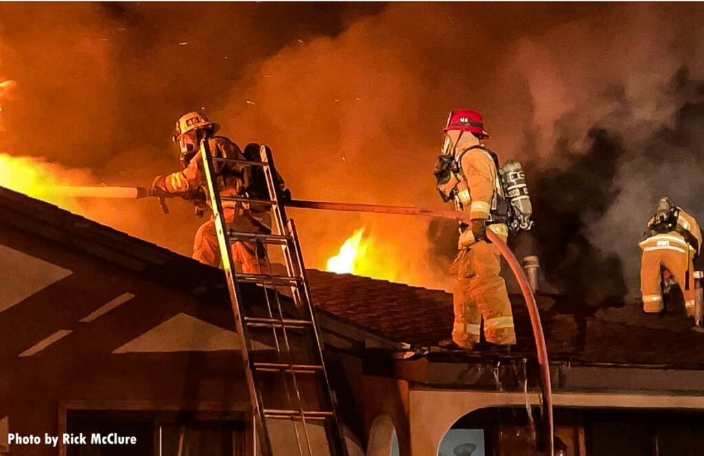 Firefighter with hose on roof of home blasting water on fire