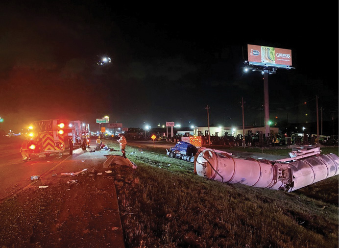 To the right of the I-10 pavement, the tank trailer lies across a ditch, broken and empty. The driver remains trapped in the tractor cab.