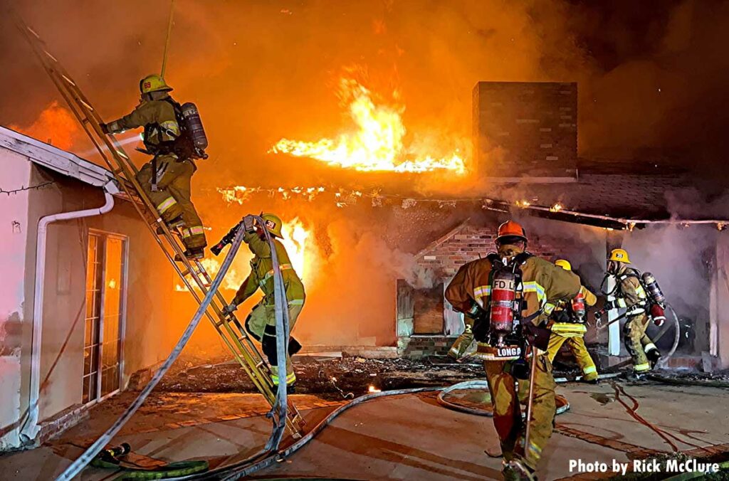 Firefighters bring a hose up a ladder as flames roar in the background