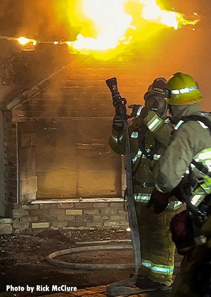 Firefighter gesture towards hoseline with flames in background