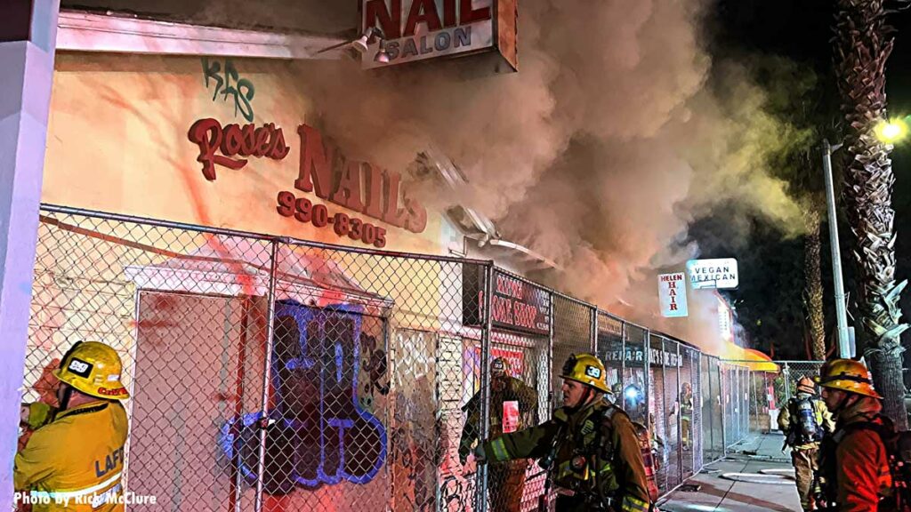 Smoke vents from row of buildings as firefighters navigate fence