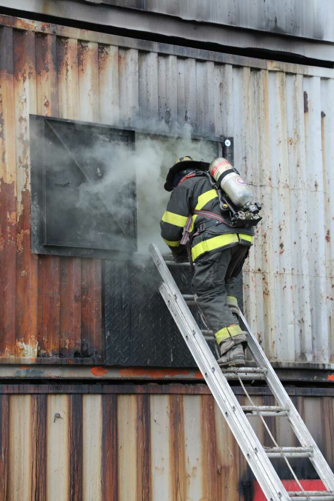 Firefighter on ladder with smoke coming from window during training FDIC 2022