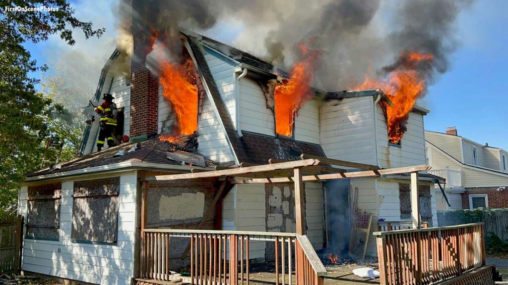 Firefighter on roof as fire shoots from three windows of house