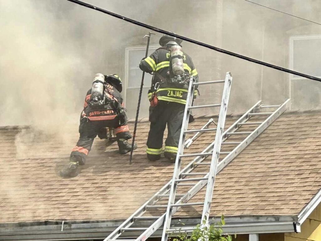 Firefighters perform vertical ventilation with one firefighter holding a hook